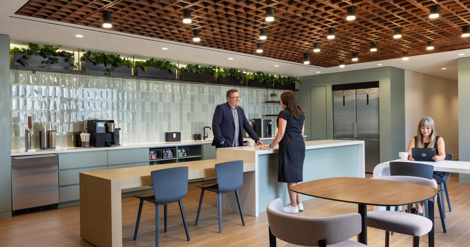 Modern kitchen and lounge area featuring a sleek, minimalist design. A wooden ceiling with geometric patterns contrasts with the smooth counter surfaces. Two professionals engage in discussion, while another individual works on a laptop at a table. Lush greenery decorates the upper shelves, enhancing the welcoming atmosphere.