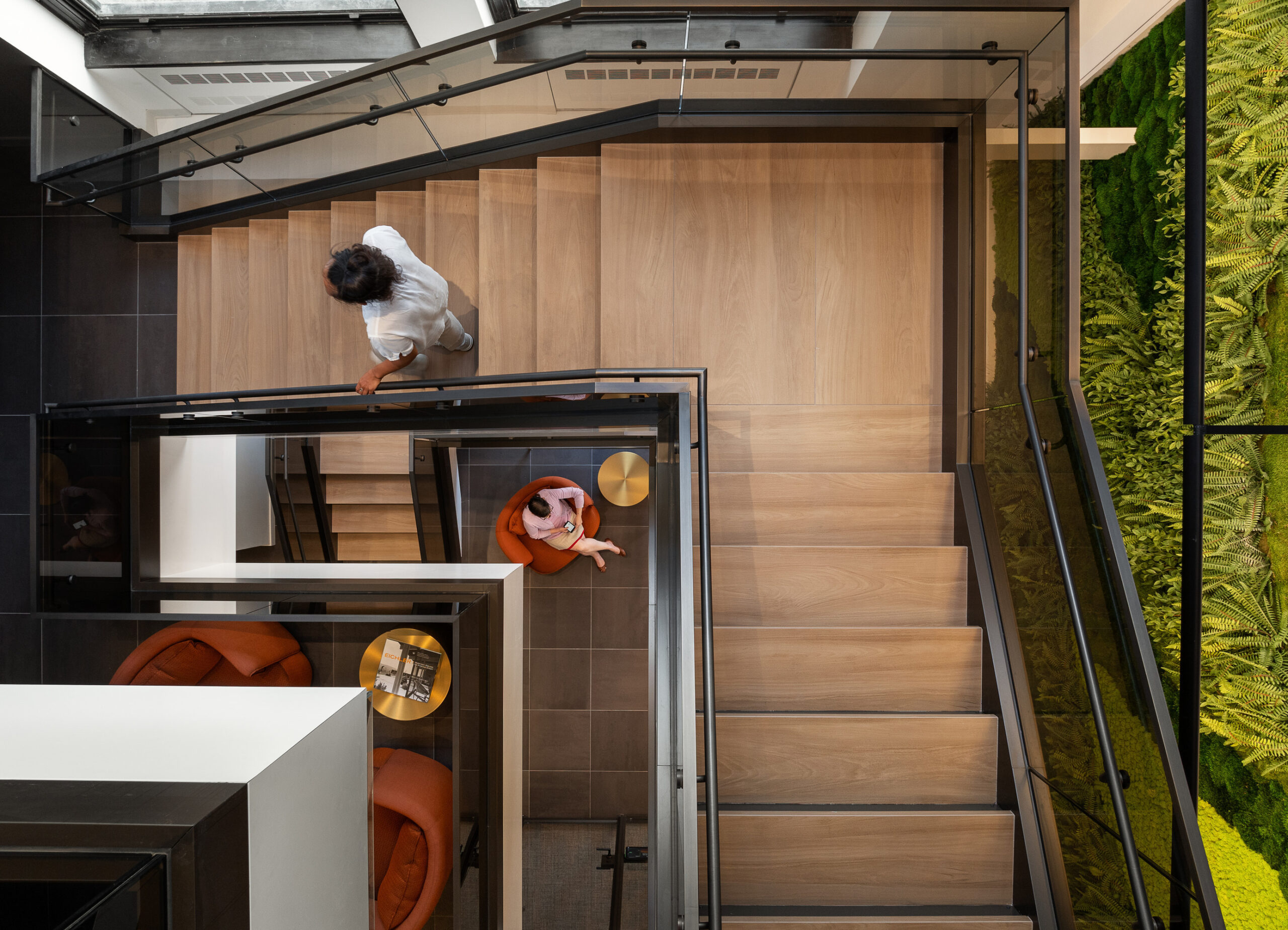 Aerial view of a modern staircase featuring sleek wooden steps and a glass railing. Below, two individuals converse in stylish orange armchairs. A vibrant vertical garden enhances the aesthetic, interplaying natural elements with contemporary design. The space emphasizes open sightlines and a harmonious blend of indoor and outdoor influences.