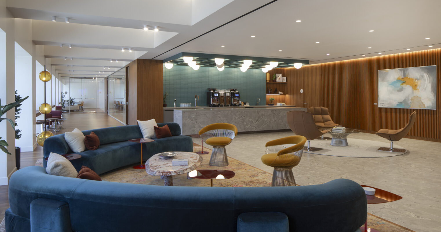 A modern lounge space featuring a curved, blue velvet sofa paired with a unique marble coffee table. The warm wood paneling contrasts with the vibrant teal backsplash behind a sleek kitchen area. Stylish lighting fixtures illuminate the room, enhancing the contemporary artistic vibe. Elegant accent chairs provide additional seating.