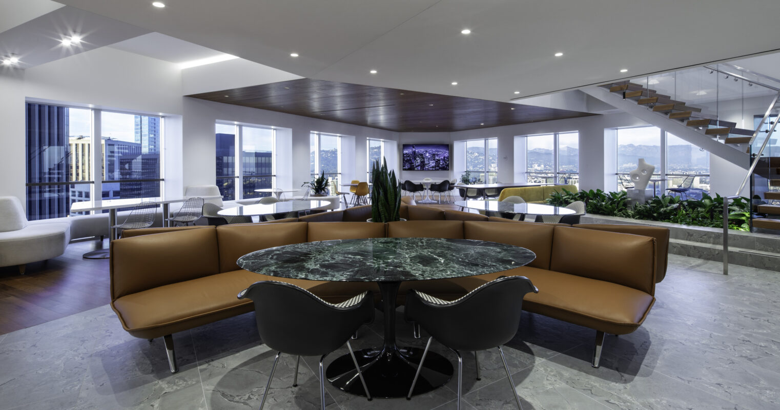 Modern office lounge featuring a circular marble-topped table surrounded by a sleek brown leather banquette and black chairs. The space is illuminated by recessed lighting and large windows, offering a panoramic view of the city skyline. Contemporary furnishings emphasize a blend of functionality and elegance, creating an inviting atmosphere.