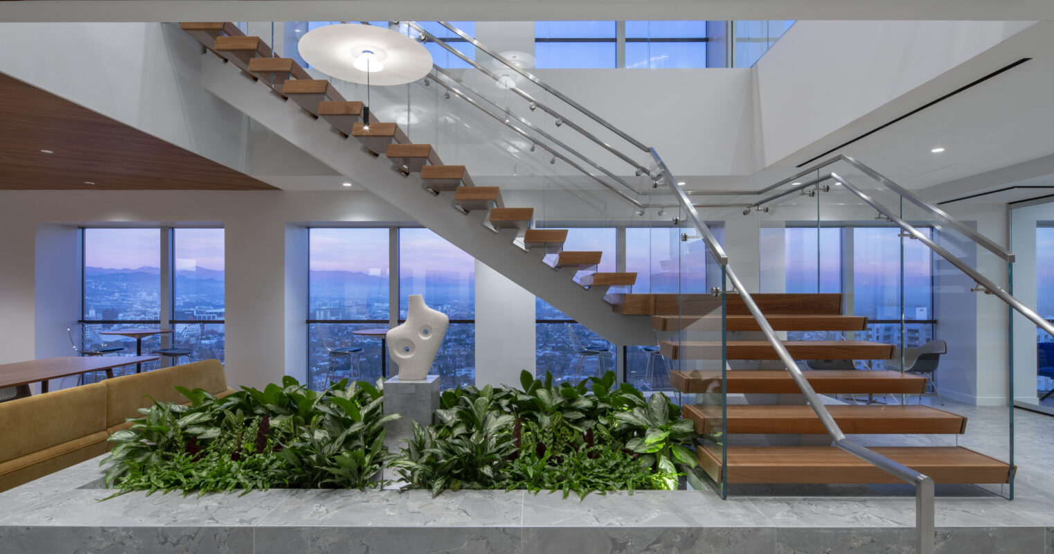 A modern interior featuring a striking staircase with wooden treads and glass railings, complemented by a sculptural piece on a marble base. Lush greenery in a stone planter adds vibrancy, while expansive windows offer panoramic views, enhancing the serene atmosphere of this stylish open space.