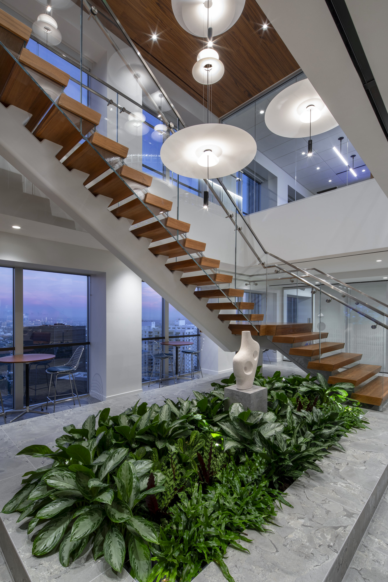 A modern staircase featuring floating wooden steps contrasts with a sleek glass railing, enhancing the contemporary aesthetic. Below, a lush arrangement of tropical plants adds a vibrant touch, while stylish pendant lights illuminate the space, highlighting the seamless integration of nature and architecture in this interior design.
