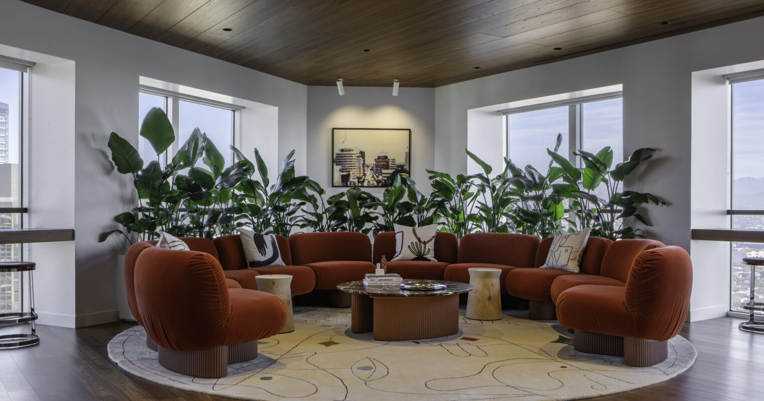 A spacious lounge area features an oversized, curved rust-colored sofa surrounded by lush indoor plants. Natural light floods the space through large windows, highlighting the rich wooden ceiling and providing a warm ambiance. A round area rug with abstract patterns anchors the seating, complemented by modern side tables.