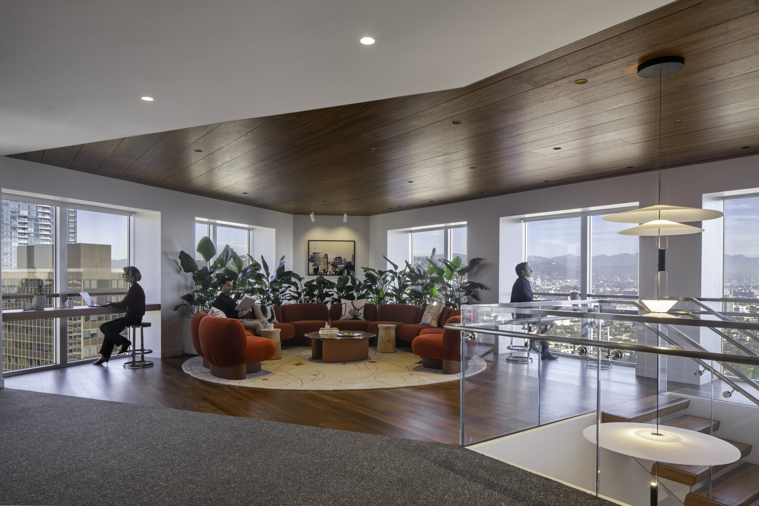 A modern office lounge featuring plush orange seating arranged around a central coffee table. Large windows allow natural light to flood the space, complemented by vibrant indoor plants. The contrast of warm wooden ceiling and sleek glass railings enhances the contemporary aesthetic while promoting a collaborative atmosphere.