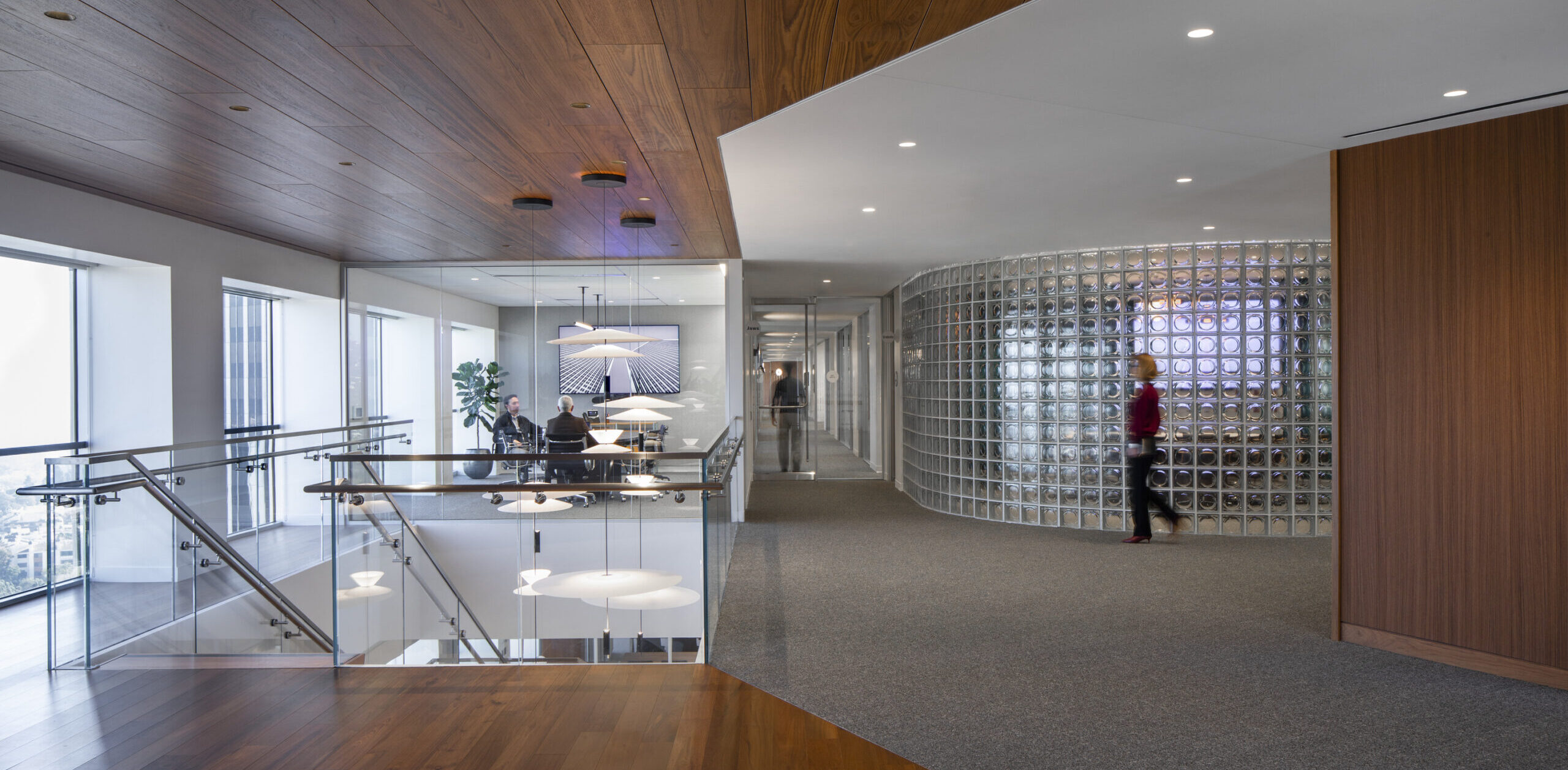 Modern office interior showcasing a blend of wood and glass materials. The sleek wooden ceiling contrasts with the transparent glass walls, fostering an open atmosphere. Curved glass partitions define workspaces, while strategically placed lighting highlights architectural features, enhancing the clean, professional aesthetic of the design.