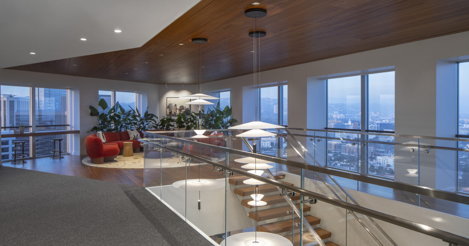 Spacious interior showcasing a modern office environment. Large windows provide panoramic city views, while lush indoor plants add a touch of nature. A sleek, wooden ceiling contrasts with the minimalist decor. The staircase, framed by glass, leads to a welcoming lounge area featuring vibrant red seating and contemporary pendant lighting.