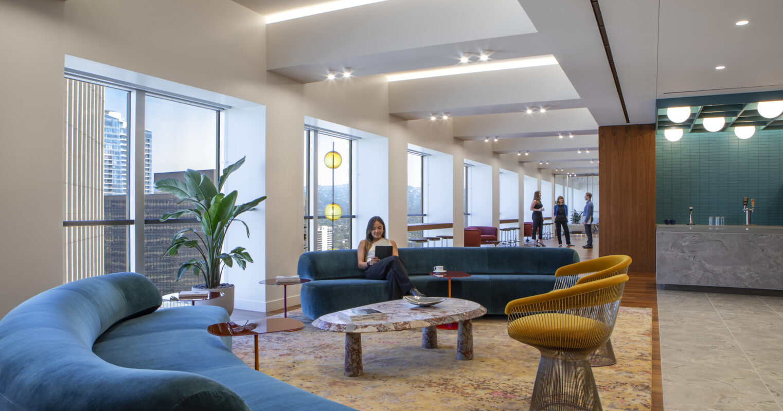 A modern lounge area features curved teal sofas complemented by a patterned area rug. Natural light floods the space through large windows, highlighting a stylish marble coffee table and vibrant accent chairs. In the background, people engage in conversation near a contemporary bar, enhancing the inviting atmosphere.
