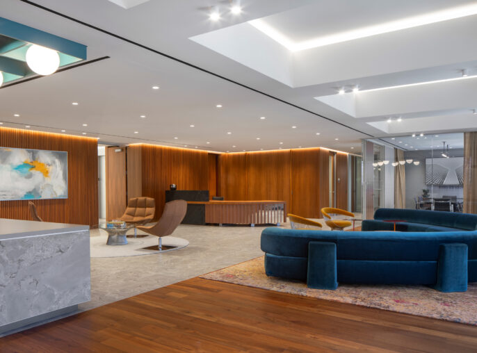 A modern open-plan interior showcasing a sophisticated lounge area, featuring a deep blue curved sofa, a stylish mid-century armchair, and a sleek marble counter. Warm wood paneling complements the contemporary design, while strategically placed lighting highlights the space's clean lines and elegant furnishings, creating an inviting atmosphere.