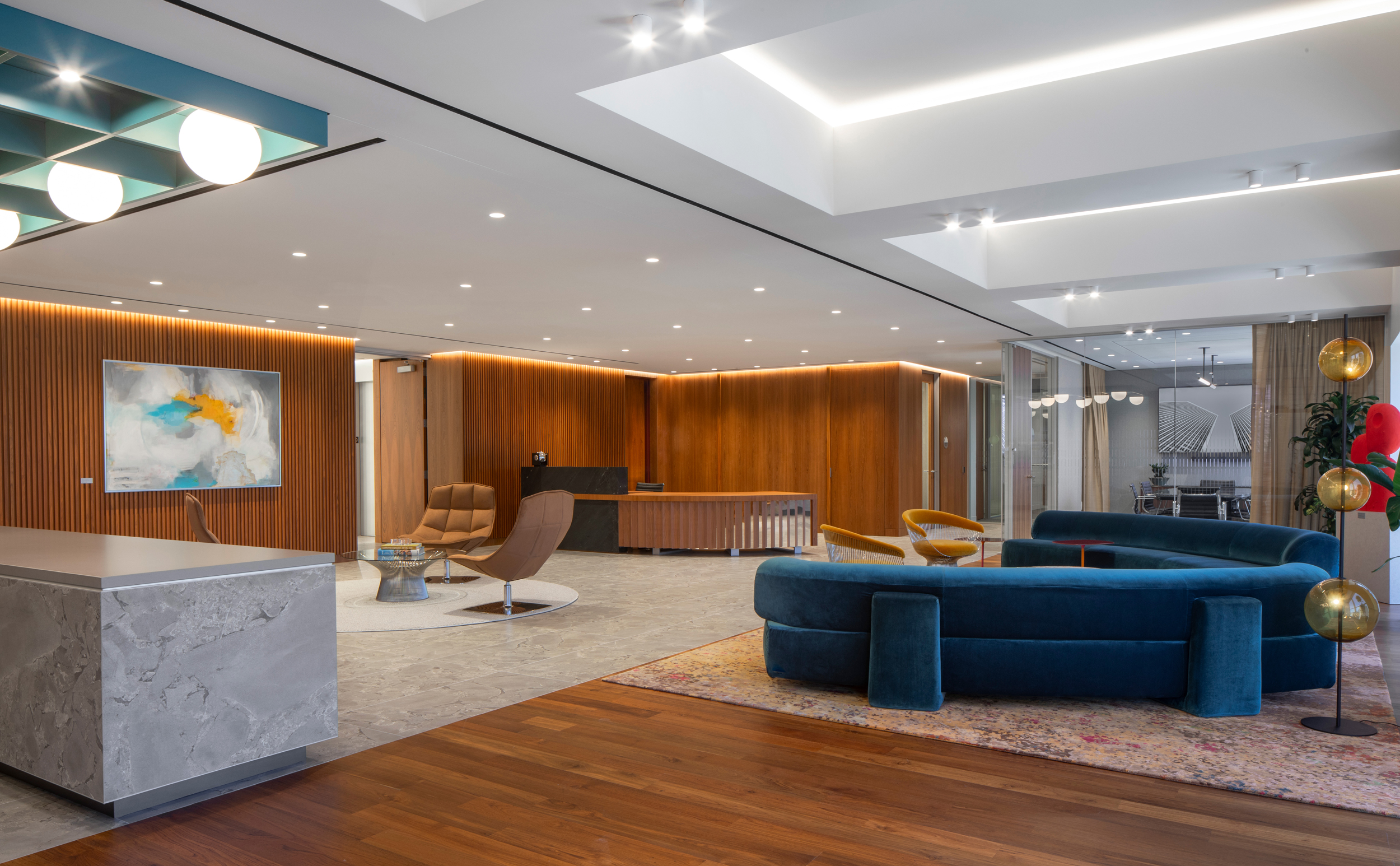 A modern open-plan interior showcasing a sophisticated lounge area, featuring a deep blue curved sofa, a stylish mid-century armchair, and a sleek marble counter. Warm wood paneling complements the contemporary design, while strategically placed lighting highlights the space's clean lines and elegant furnishings, creating an inviting atmosphere.