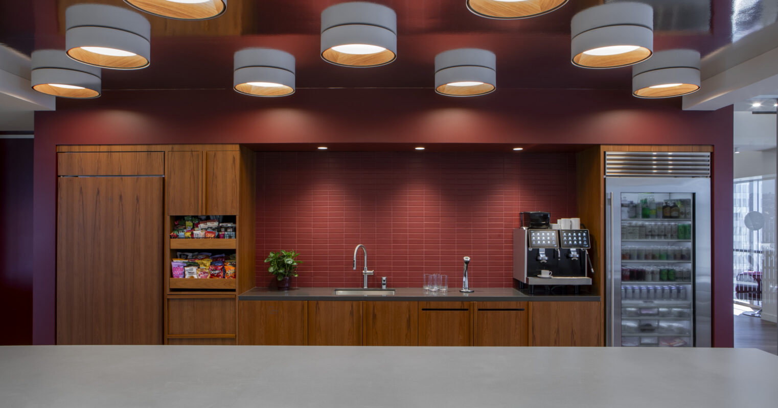This modern kitchen features rich walnut cabinetry and a dark quartz countertop, complemented by deep red walls and a vibrant backsplash. Elegant pendant lights illuminate the space, enhancing the sleek design. A well-organized coffee station and an array of snacks add functionality and warmth to the environment.