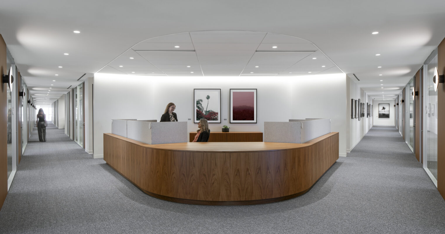 A modern office reception area featuring a smooth, curved walnut front desk. Soft, ambient lighting complements the minimalistic design. Framed artwork adorns the walls, providing a touch of sophistication. The sleek, open layout encourages collaboration, while muted gray carpeting enhances the tranquil atmosphere throughout the corridor.