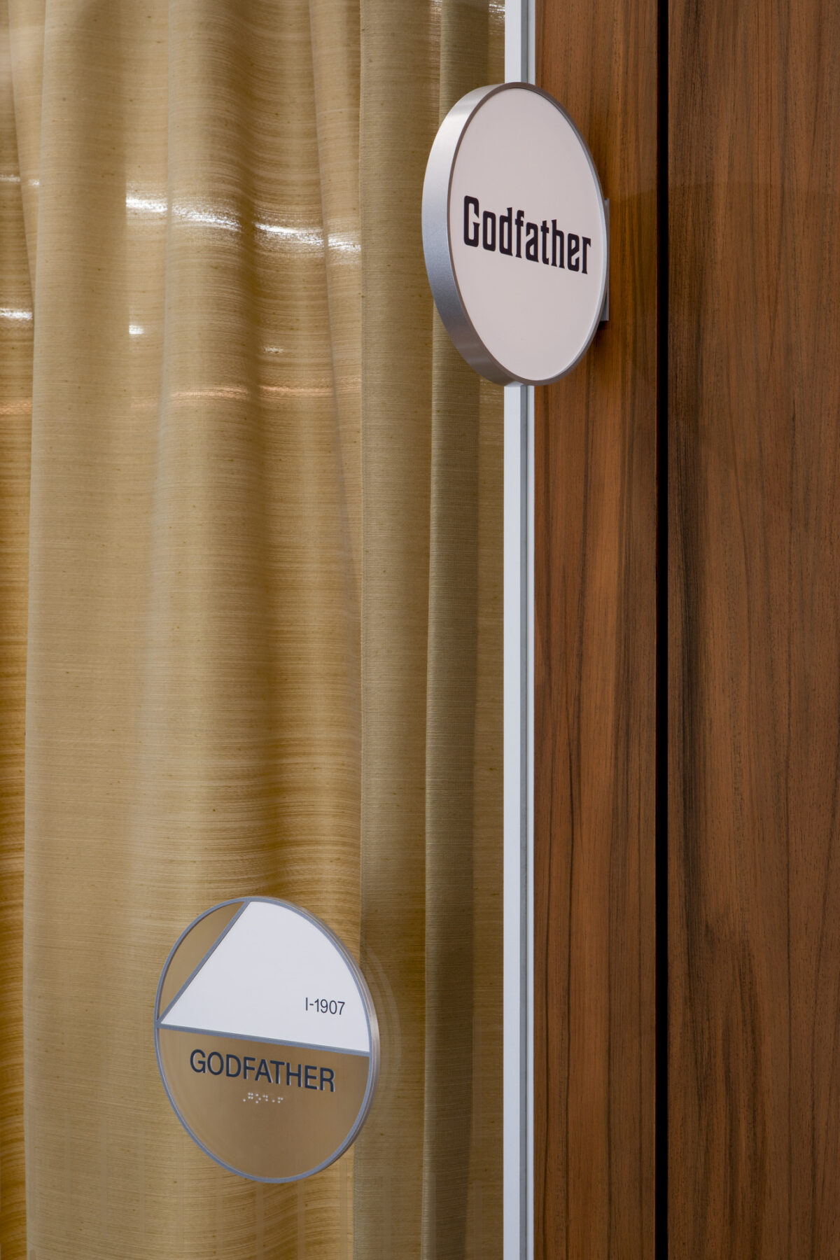 Elegant signage complements the warm wood paneling and soft fabric drapery in this interior space. The circular nameplates use a modern font, contrasting with the sophisticated textures, while subtle lighting enhances the inviting atmosphere. This design exemplifies the balance between functionality and aesthetic appeal in contemporary interiors.