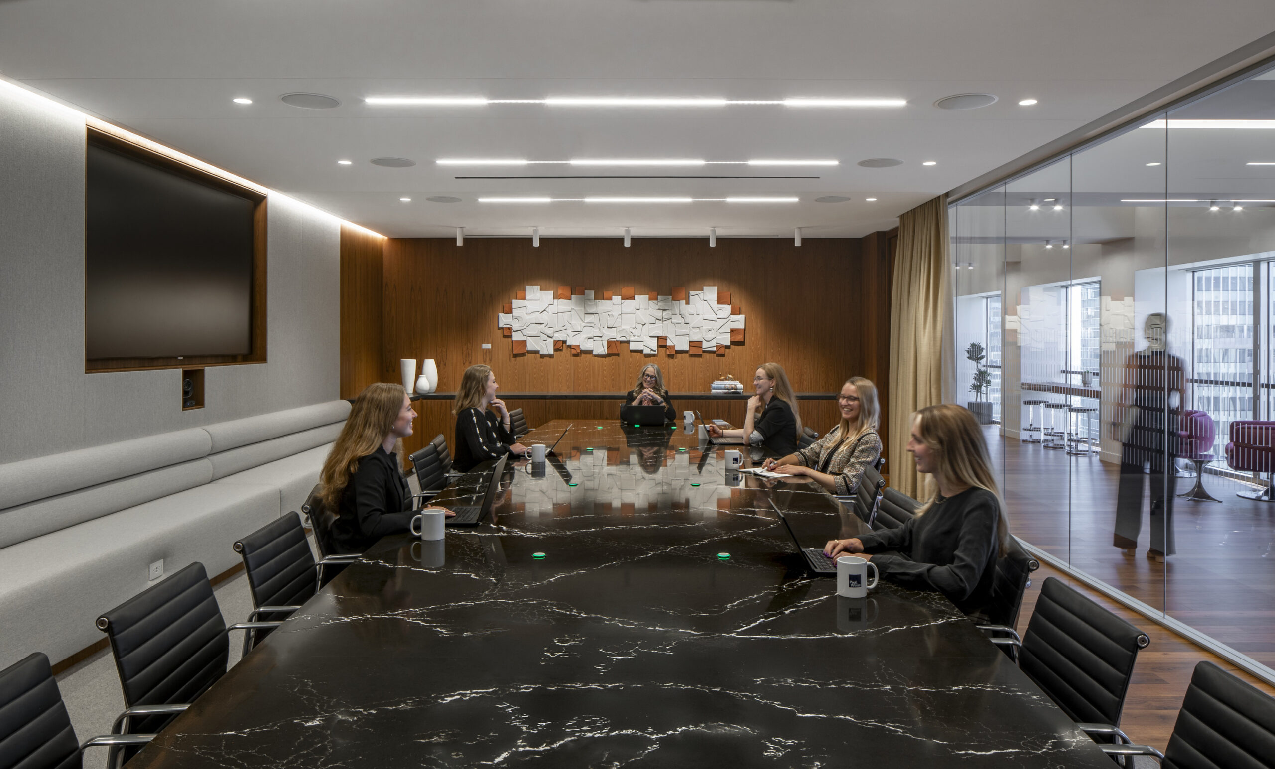 A modern conference room featuring a sleek, elongated black marble table surrounded by ergonomic leather chairs. On one wall, a textured wood panel provides warmth, contrasting with the contemporary design. A large screen and ambient lighting enhance the professional atmosphere, while individuals engage in a meeting, reflecting collaboration.