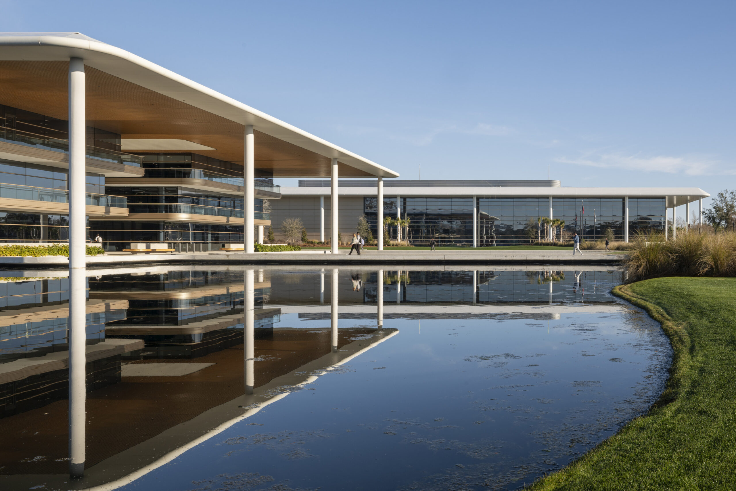 Modern corporate campus featuring expansive glass façades, sweeping rooflines, and white columns overlooking a reflective pond, with landscaped grounds and pedestrian walkways under a clear blue sky.