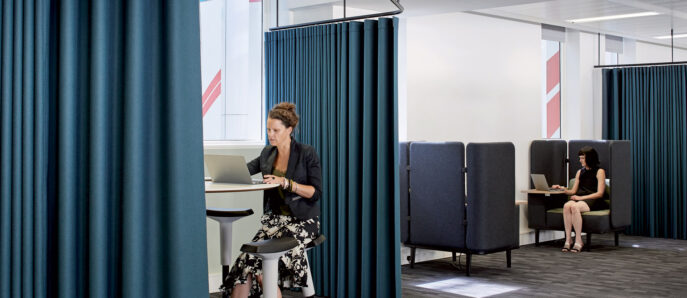 Modern office space with deep blue privacy curtains dividing workstations, featuring individuals focused on portable devices. The design balances open layouts with seclusion, coupled with minimalist high stools and task lighting to create an airy yet functional workspace.