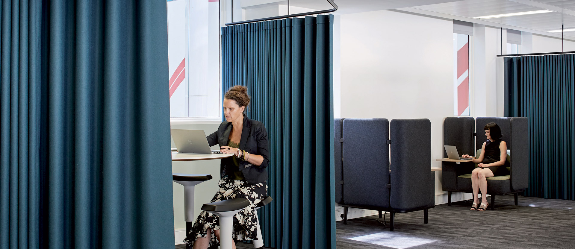 Modern office space with deep blue privacy curtains dividing workstations, featuring individuals focused on portable devices. The design balances open layouts with seclusion, coupled with minimalist high stools and task lighting to create an airy yet functional workspace.