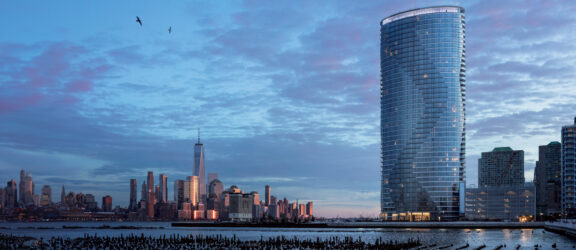 A modern curved glass building on the river in Jersey City, set against the iconic New York City skyline in the background. The partly cloudy sky adds to the serene atmosphere, and wooden posts protrude from calm waters in the foreground.