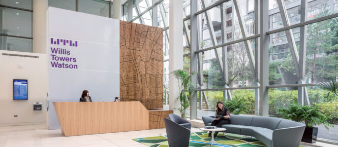 Spacious atrium lobby featuring high ceilings with geometric skylights, a large scale wood-paneled reception desk, sleek gray sofas on patterned area rugs, and airy greenery, embodying modern corporate design.