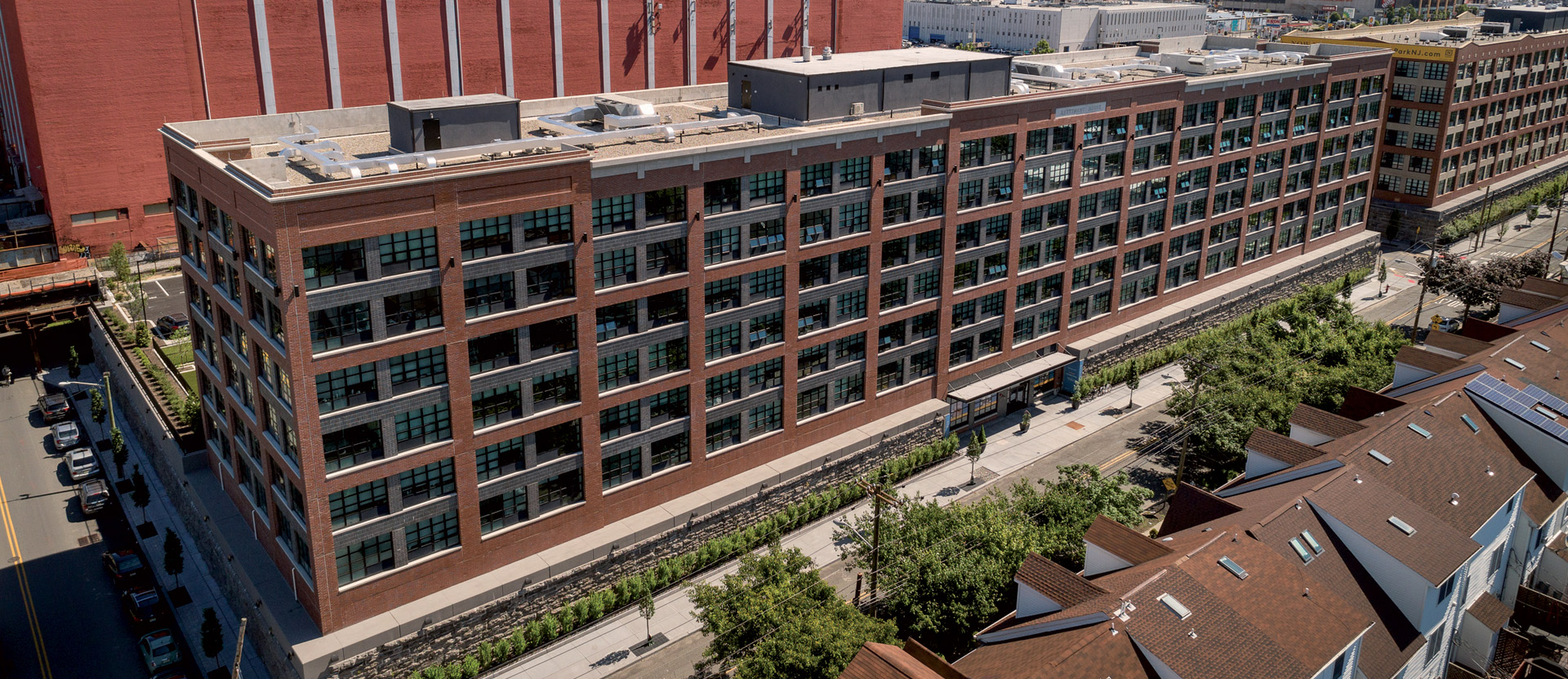 Modern urban apartment building featuring a blend of brick and large windows, surrounded by a developing neighborhood with a skyline backdrop.