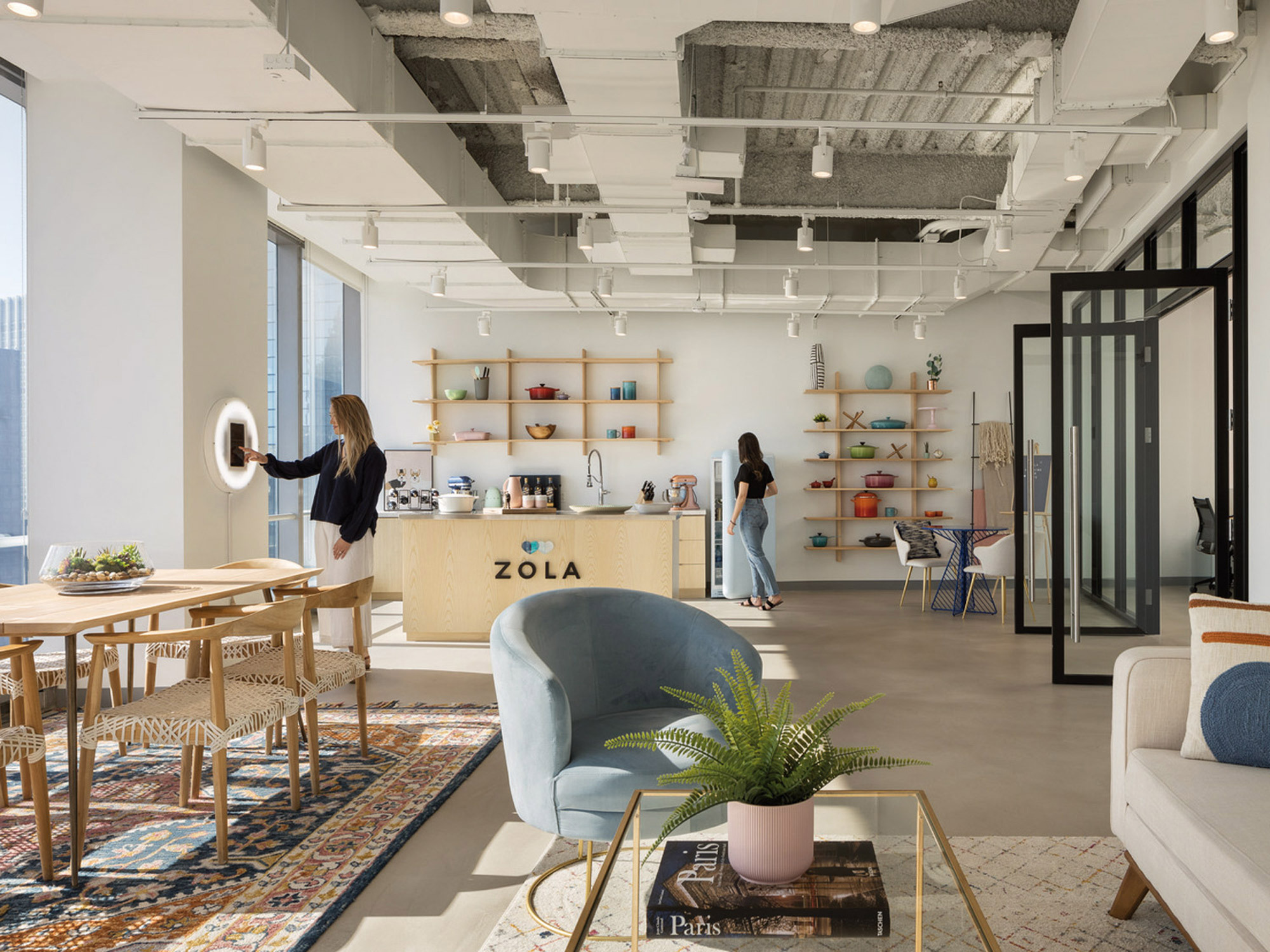Exposed concrete ceilings and ductwork juxtapose the modern elegance of an open-plan interior. Natural light floods through large windows, illuminating an eclectic mix of textures and colors. A central plywood shelving unit, branded with 'ZOLA', displays an array of curated kitchenware, infusing a sense of homeliness. Woven area rugs overlay the polished concrete floor, anchoring assorted furniture pieces that articulate