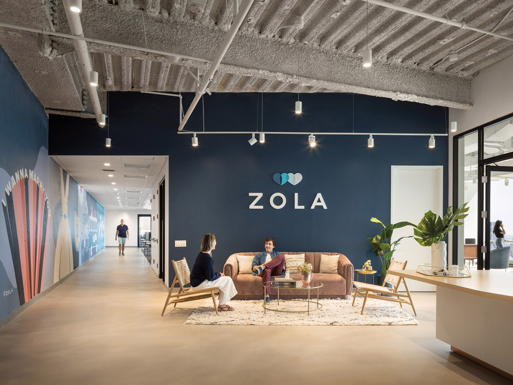 Spacious corporate interior features an exposed concrete ceiling with suspended linear lighting, enhancing a modern industrial aesthetic. Rich navy walls contrast with warm wooden accents and a bespoke wall mural, while contemporary furniture provides casual seating areas for collaboration. Strategic placement of indoor plants introduces a natural, calming element within the workspace.