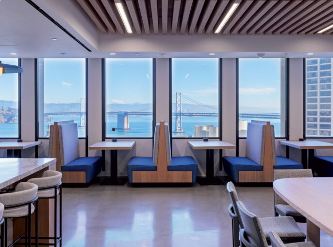 Modern office break room overlooking a bridge through expansive windows, featuring wooden slat ceiling accents, sleek pendant lighting, and two-tone banquette seating. Neutral flooring complements the space's minimalist aesthetic.