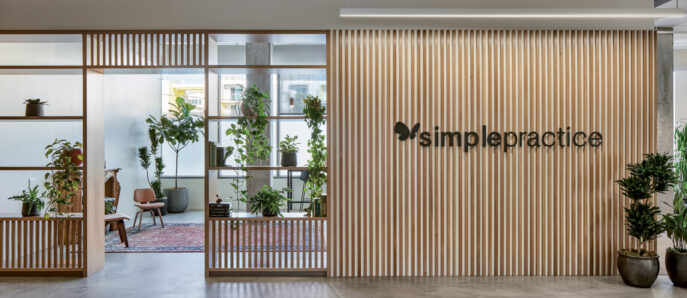 Modern office lobby featuring vertical wooden slats creating a semi-transparent partition, accented with potted green plants. Elegantly furnished with minimalistic seating and a warm, neutral color palette complemented by ambient lighting, promoting an inviting and contemporary workspace atmosphere.