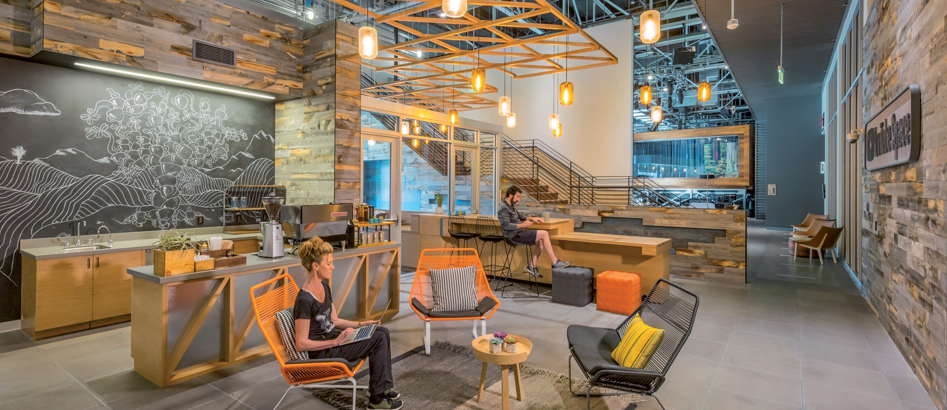 Exposed ceilings and industrial lighting fixtures give this modern lounge area a spacious yet inviting ambiance. Wooden accents, including reclaimed wood wall cladding and ceiling beams, harmonize with the metalwork. Geometric furniture pieces, in a mix of vibrant and muted tones, provide functional seating while contributing to the dynamic aesthetic. An expansive chalk mural acts as a focal point, softening the industrial elements with organic lines.