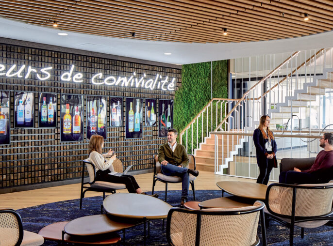 Contemporary office lounge with wooden slatted ceiling curving downward, incorporating ambient lighting. Dark marble floor hosts a cluster of elegant rattan chairs on a circular navy rug. A living green wall and illuminated brand motto enhance communal aesthetic. People converse and work on laptops, suggesting dynamic use.