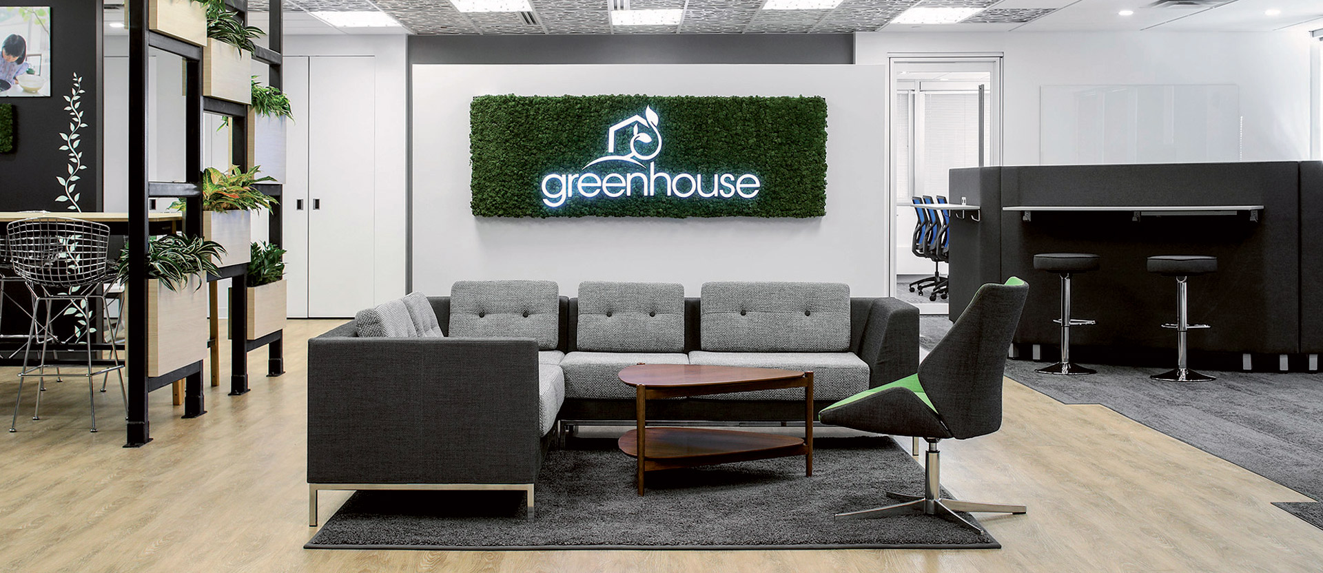 Modern office lobby featuring a neutral palette with grey sofas, a central wooden coffee table, and a moss wall displaying the "greenhouse" logo. Strategically placed greenery complements the natural aesthetic, while subtle lighting accentuates the space's clean lines and contemporary design.