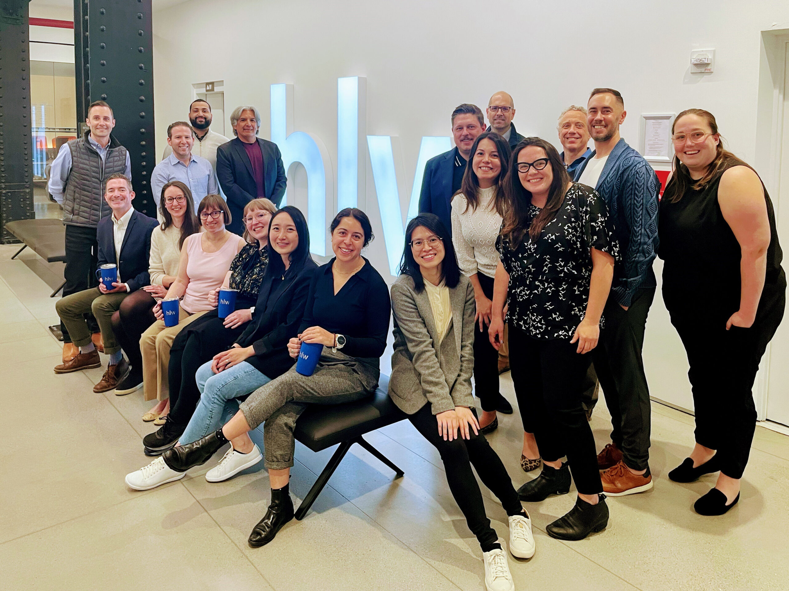 A group of smiling people posing for a photo in an office lobby with a large, illuminated "HLW" sign in the background.