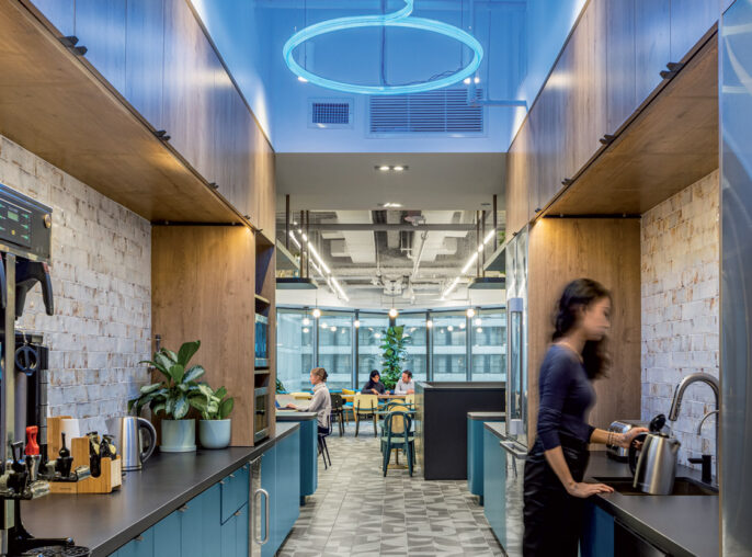 Modern kitchen area in a commercial space featuring sleek, dark wood cabinetry and LED circular lighting fixtures overhead. Patterned gray and white flooring harmonizes with the cool blue accents, while stainless steel appliances offer a contemporary touch. The open design invites interaction in an adjacent communal area.