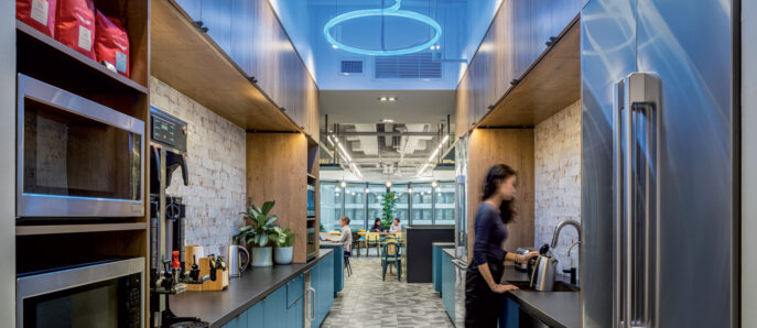 Modern kitchen area in a commercial space featuring sleek, dark wood cabinetry and LED circular lighting fixtures overhead. Patterned gray and white flooring harmonizes with the cool blue accents, while stainless steel appliances offer a contemporary touch. The open design invites interaction in an adjacent communal area.