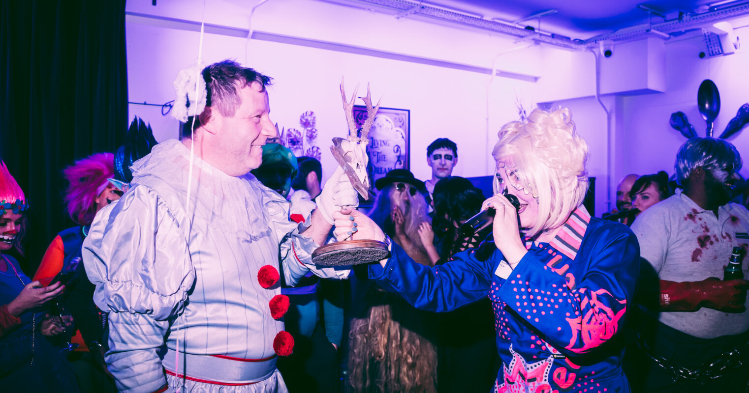 A humorous mock award ceremony with one person in a baby costume receiving a trophy, amidst a backdrop of varied costumes and fun.