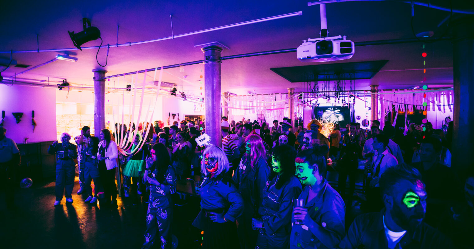 A vibrant party scene with attendees in various costumes illuminated by purple lights.