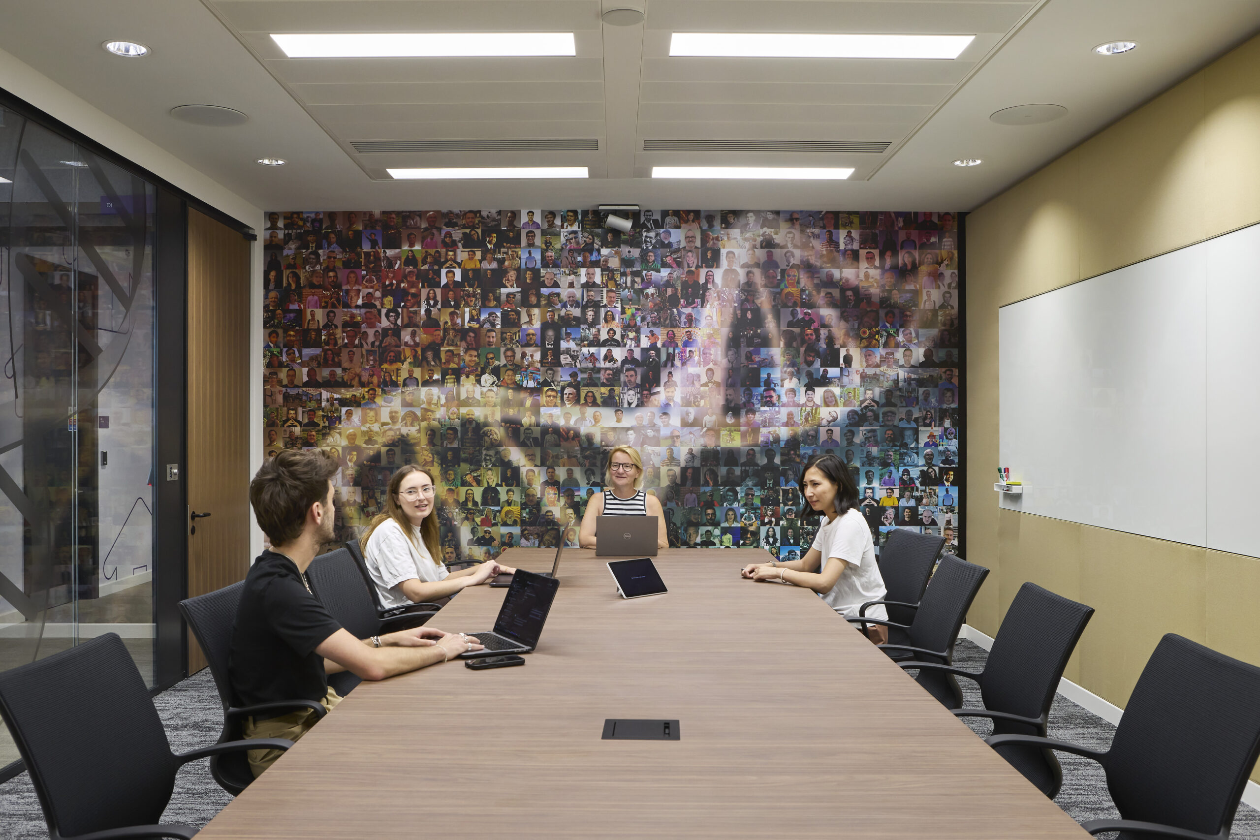 Modern conference room centered on a long wood meeting table with laptops and ergonomic black chairs. A large, colorful mosaic mural of diverse portraits forms a distinctive focal wall, complemented by whiteboards, glazed partitions, warm wood detailing, acoustic carpet, and evenly distributed recessed ceiling lighting.