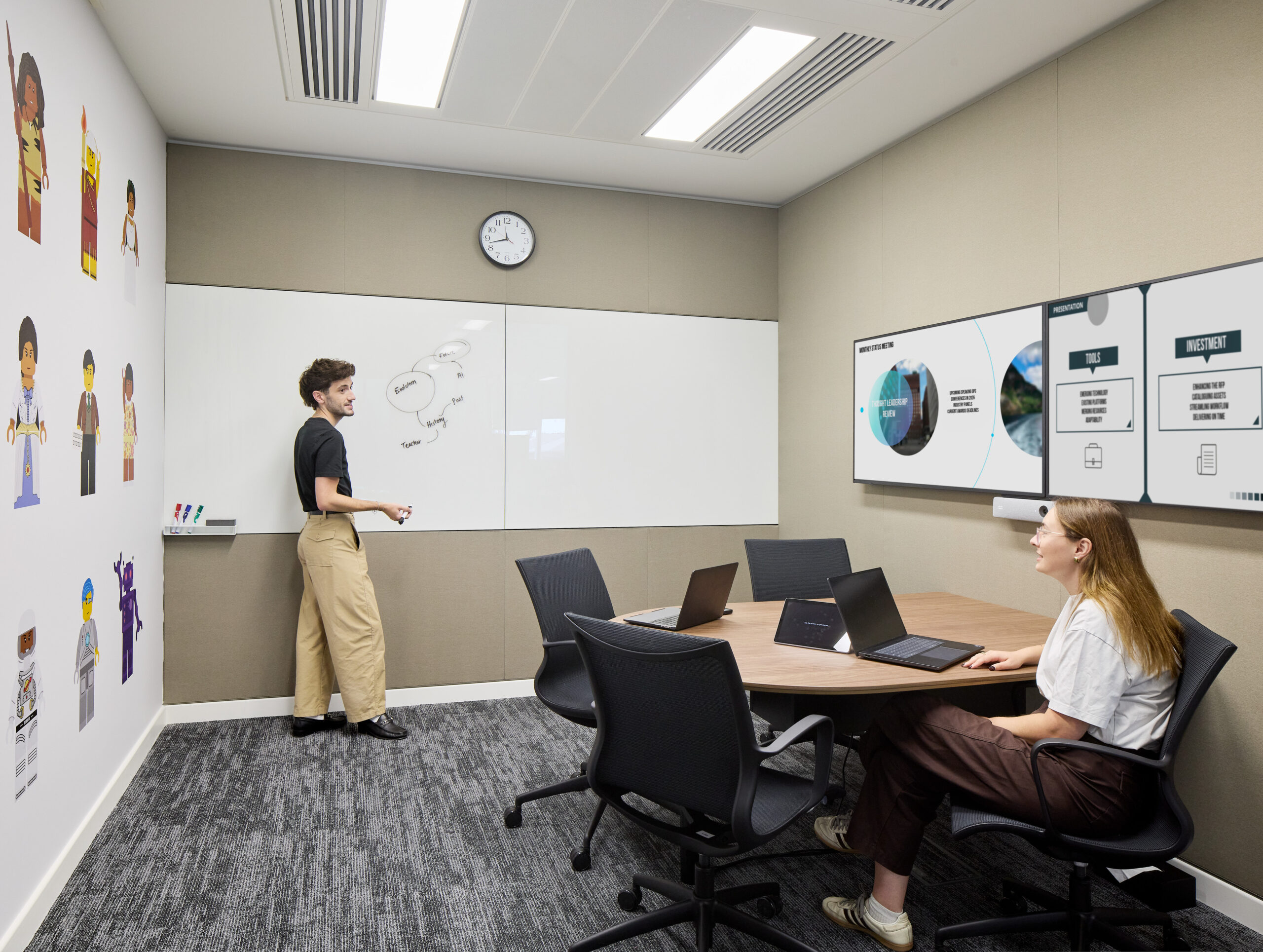 Small collaborative meeting room with a rounded wood table, ergonomic task chairs, wall-mounted presentation displays, and a full-width whiteboard marked with handwritten diagrams. Neutral acoustic wall panels and carpet provide visual calm, while colorful illustrated figures on the adjacent wall introduce a playful workplace identity.