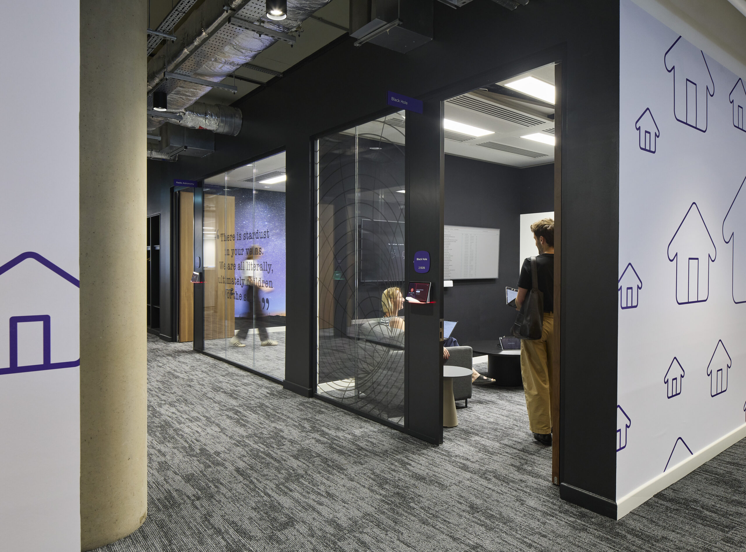 Workplace corridor lined with enclosed glass meeting rooms framed in black, dark walls, and purple room signage. One glass partition carries a large quotation, while adjacent white walls feature repeating purple house icons. Exposed ductwork, acoustic carpet, and warm timber doors combine technical and graphic design elements.