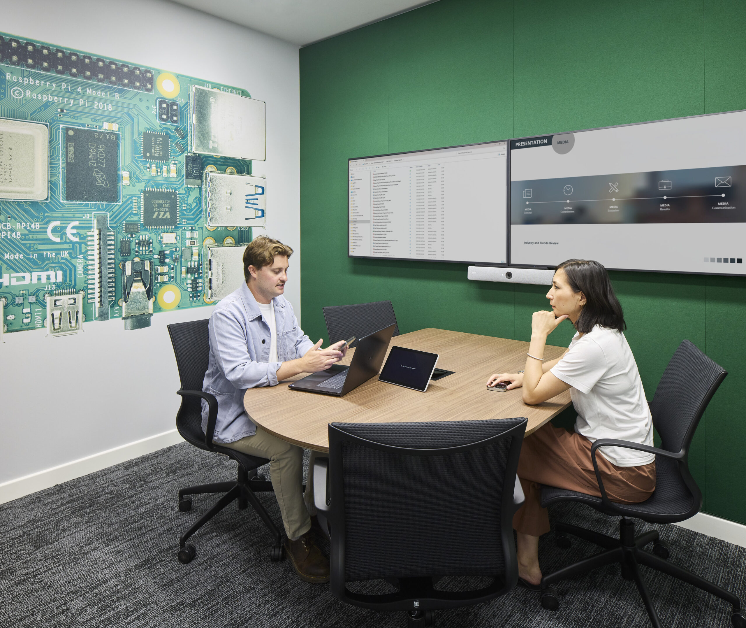 Compact meeting room with a rounded wood table, ergonomic black chairs, laptops, and dual wall-mounted digital displays. A vivid oversized circuit-board mural references technology, while a deep green acoustic wall adds contrast and warmth. Integrated video-conferencing equipment supports hybrid collaboration.