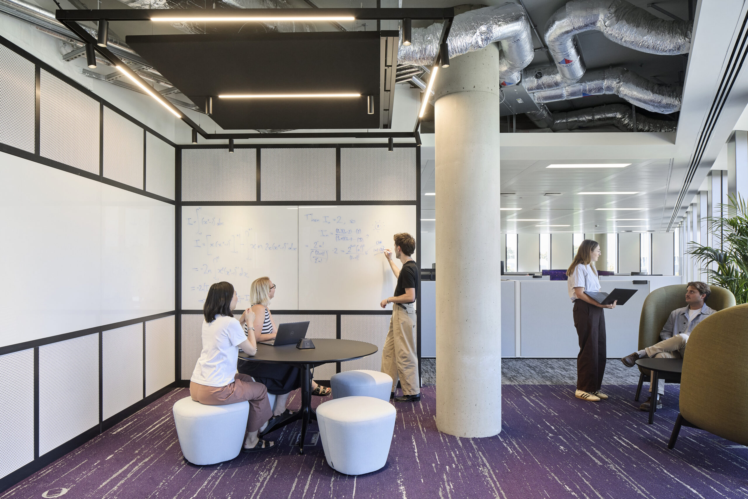 Open-plan collaboration area combining a framed whiteboard room with black metal and perforated acoustic panels, a round table, and sculptural white stools. Beyond the concrete column, lounge seating supports informal meetings. Purple patterned carpet, exposed services, and linear lighting reinforce the contemporary workplace character.