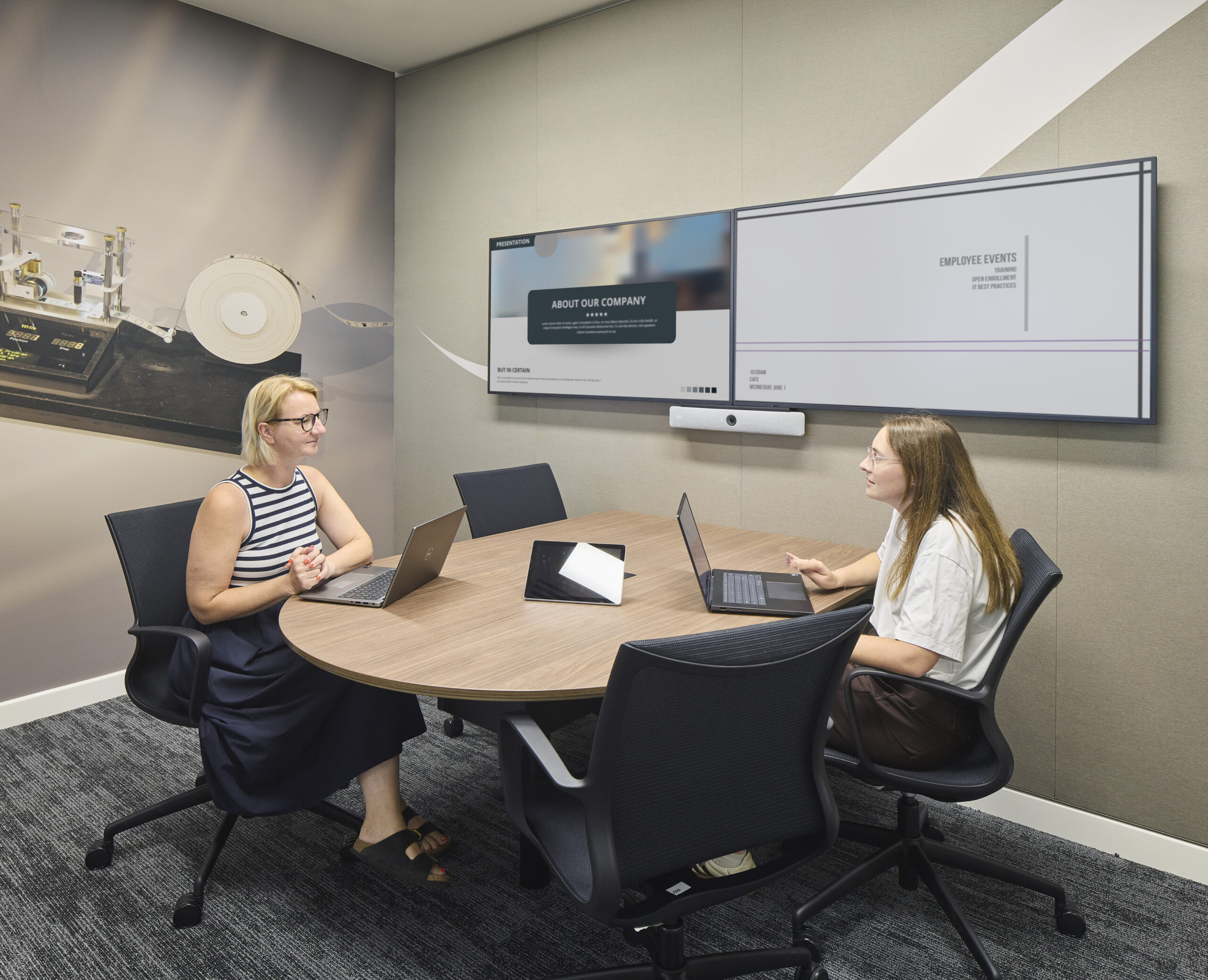 Intimate meeting room with a rounded wood table, laptops, ergonomic black chairs, and a wide digital presentation screen displaying company content. Neutral acoustic wall panels create a calm backdrop, while a large photographic mural of technical equipment adds visual depth and reinforces the organization’s technology-focused identity.