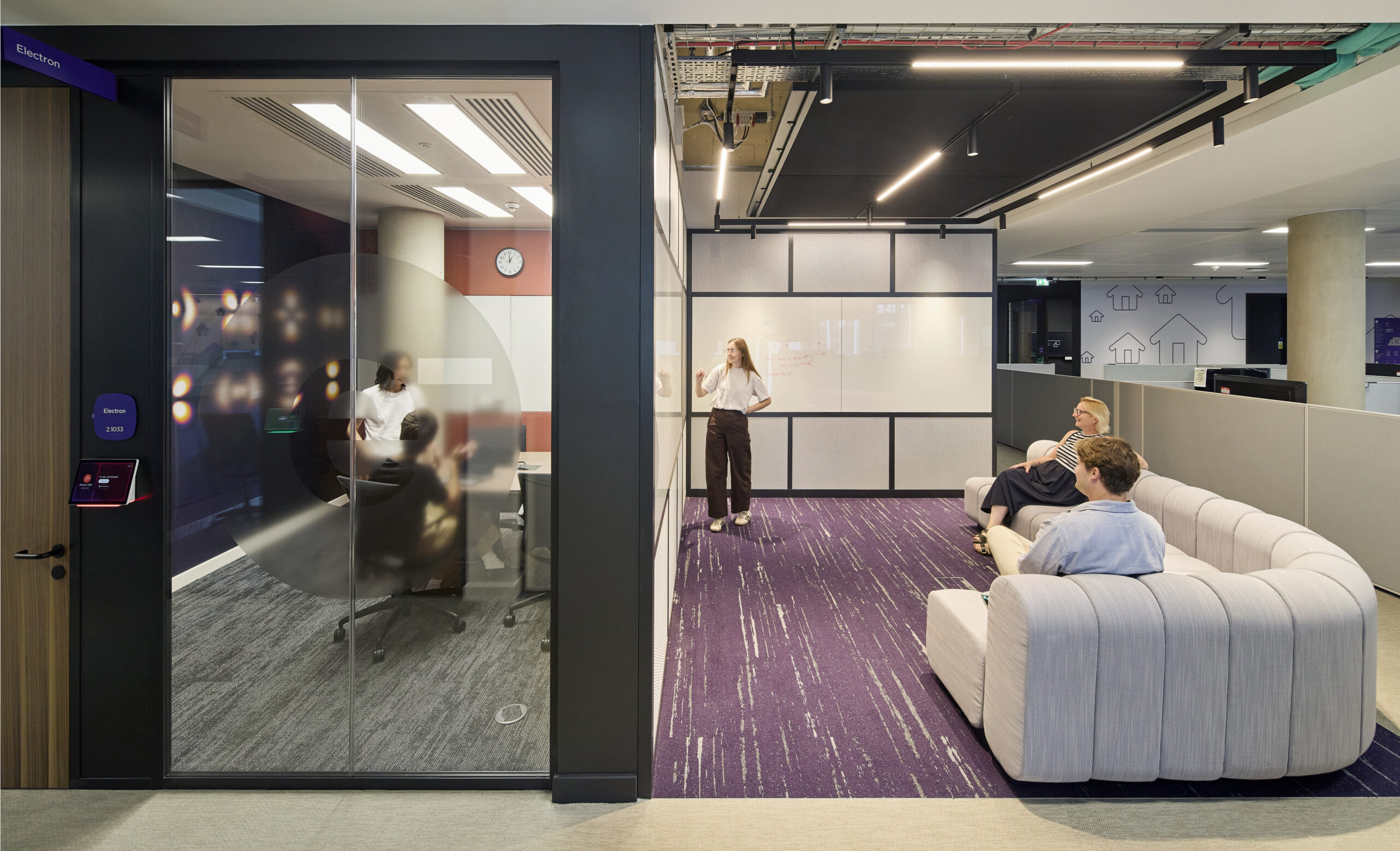 Contemporary workplace lounge beside a glazed meeting room, featuring a sculptural pale upholstered sofa, purple patterned carpet, and informal seating. Black-framed whiteboard partitions, exposed ceiling services, integrated linear lighting, and graphic house motifs create a flexible, visually distinctive environment for collaboration and casual work.