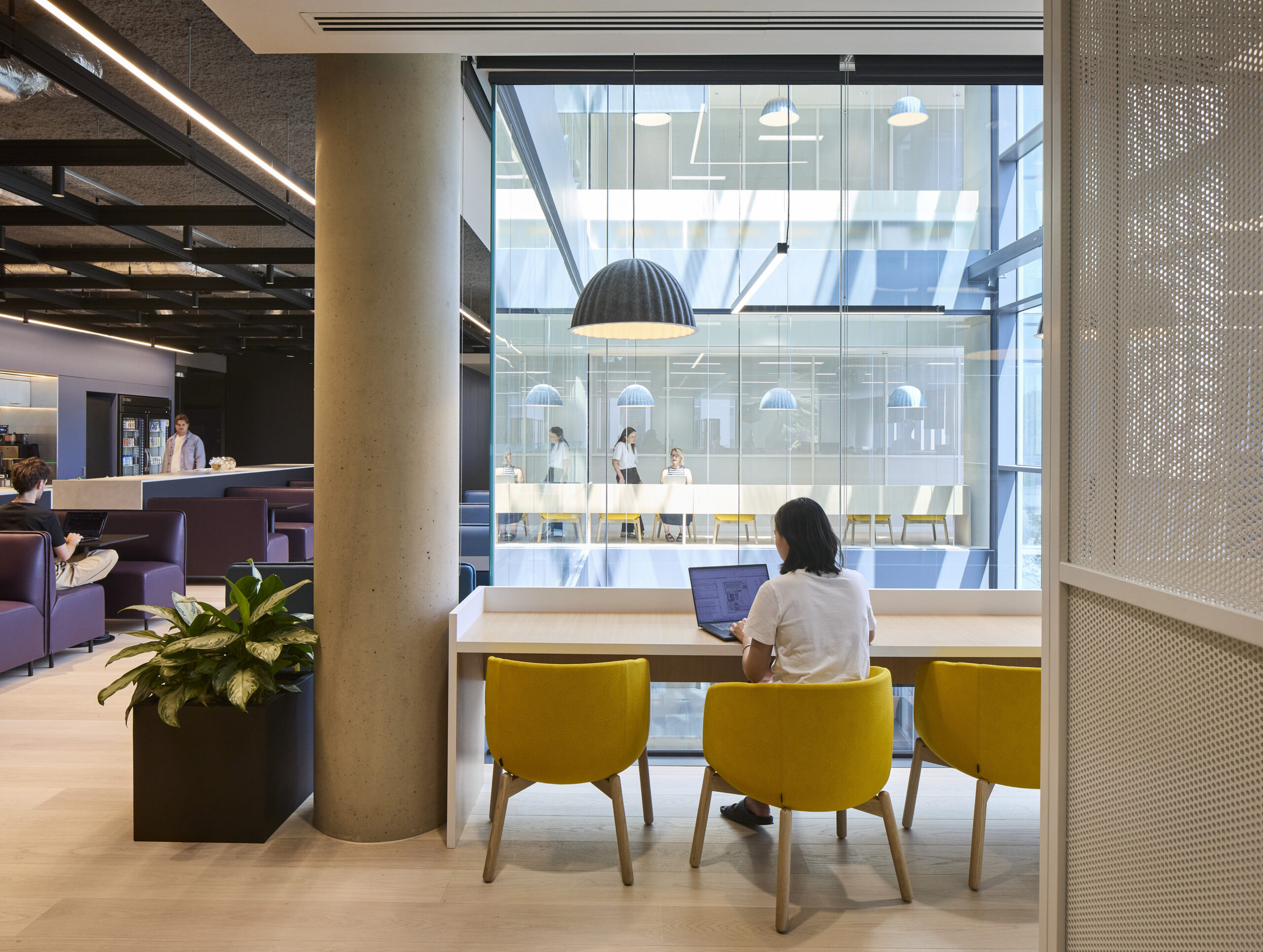 Bright workplace café overlooking a glazed internal atrium, with a built-in timber counter, yellow upholstered chairs, and a worker using a laptop. Black pendant lights, exposed ceiling elements, indoor planting, pale timber flooring, and adjacent lounge seating combine hospitality with flexible individual work settings.