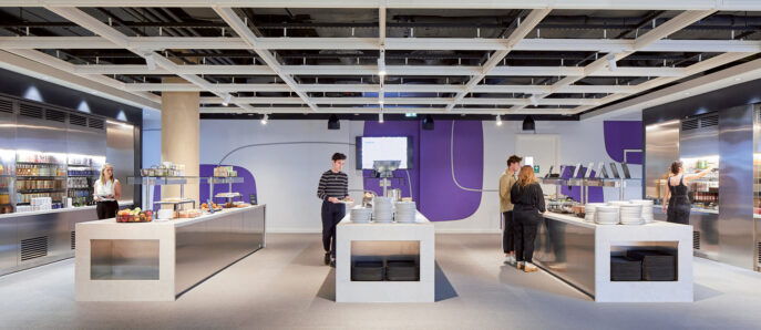 Modern office cafeteria with exposed ceiling, featuring sleek white furniture and vibrant purple accent walls. The space is well-lit, promoting a clean and energizing atmosphere for break-time refreshment.