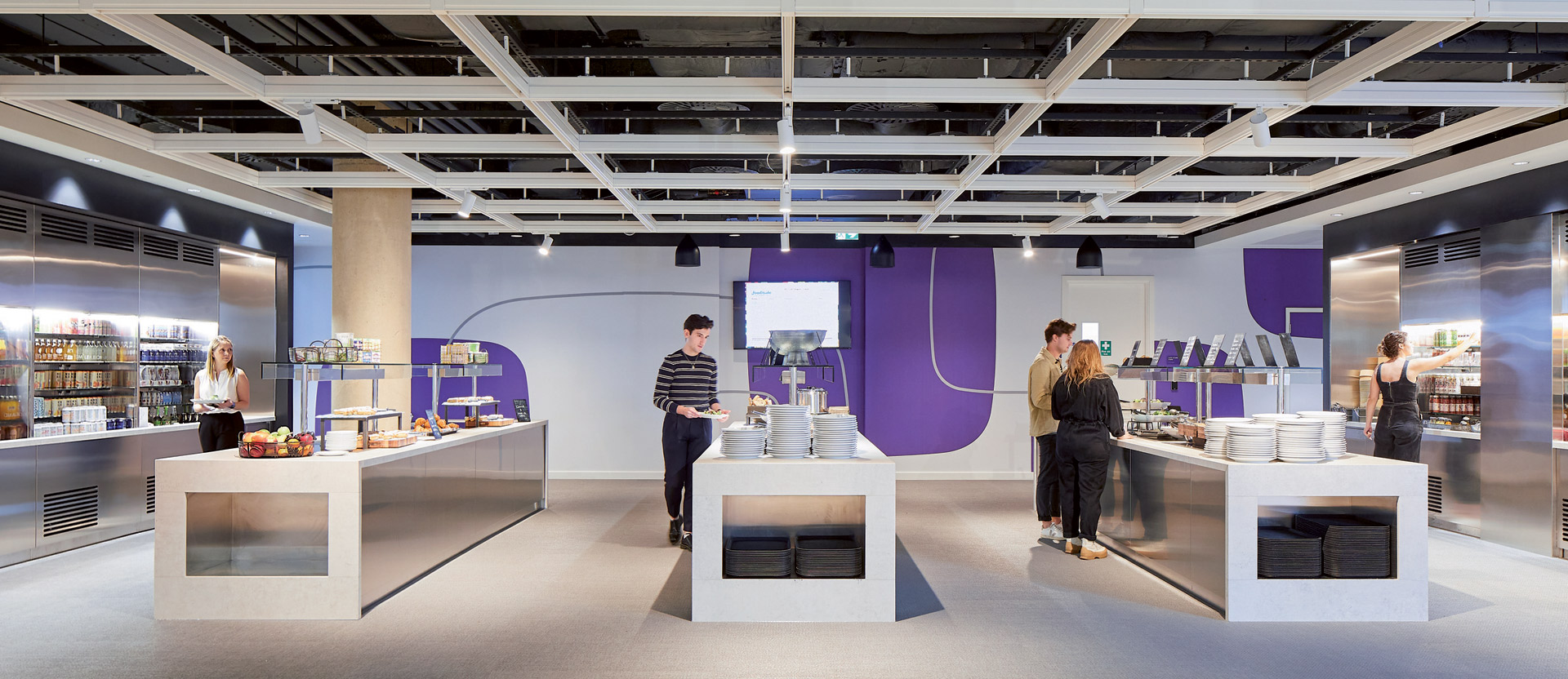 Modern office cafeteria with exposed ceiling, featuring sleek white furniture and vibrant purple accent walls. The space is well-lit, promoting a clean and energizing atmosphere for break-time refreshment.