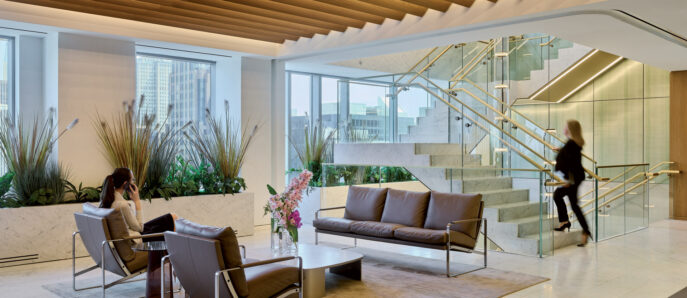 A modern office lobby featuring a recessed ceiling with wooden slats, marble stairs with glass railings, and seating areas with cityscape views.