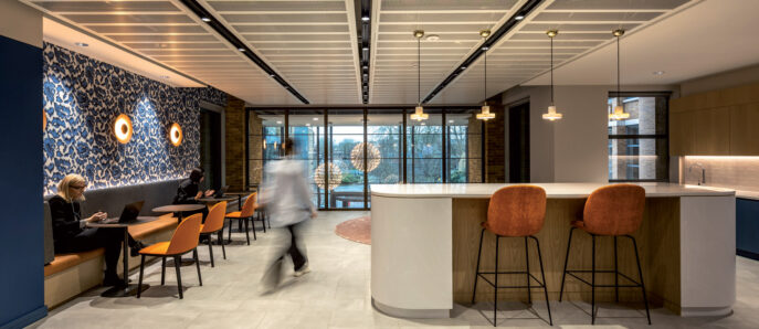 Modern office break room featuring a dynamic balance of soft seating and high-top bar stools, with contrasting textures including a sleek wood finish kitchenette, blue patterned accent wall, and exposed linear ceiling elements, all illuminated by natural light from expansive windows.