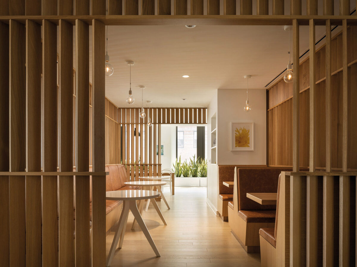 Warm-toned wooden slats create a semi-open partition in a well-lit, modern interior, framing the view into an inviting space with minimalist furniture and pendant lighting, accented by a splash of color from abstract wall art.