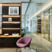 A cozy corner in an office with a modern shelf illuminated by warm lights, a purple chair, and framed artwork.