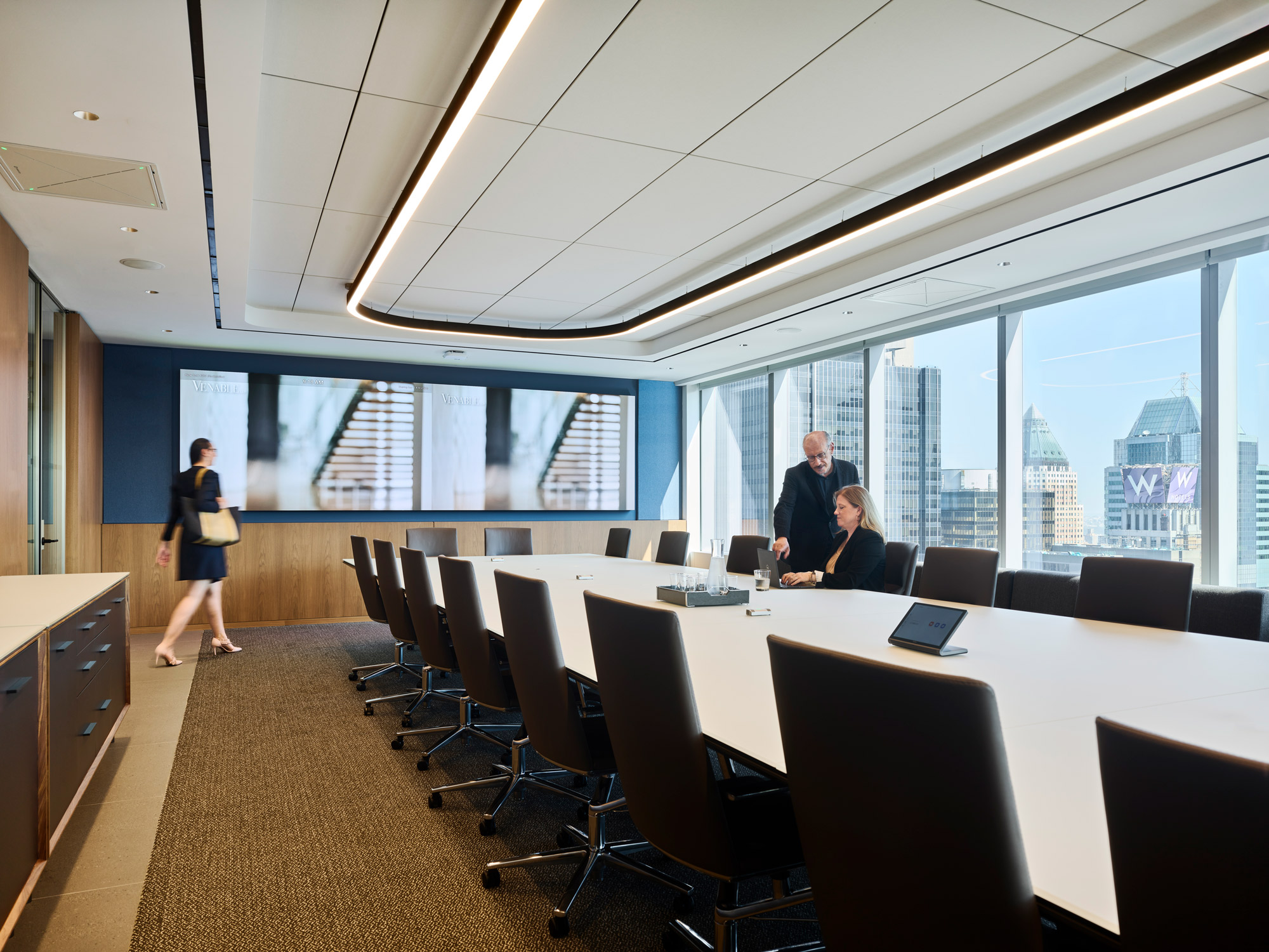 A boardroom with a large conference table, overhead linear lighting, and floor-to-ceiling windows showcasing city views.